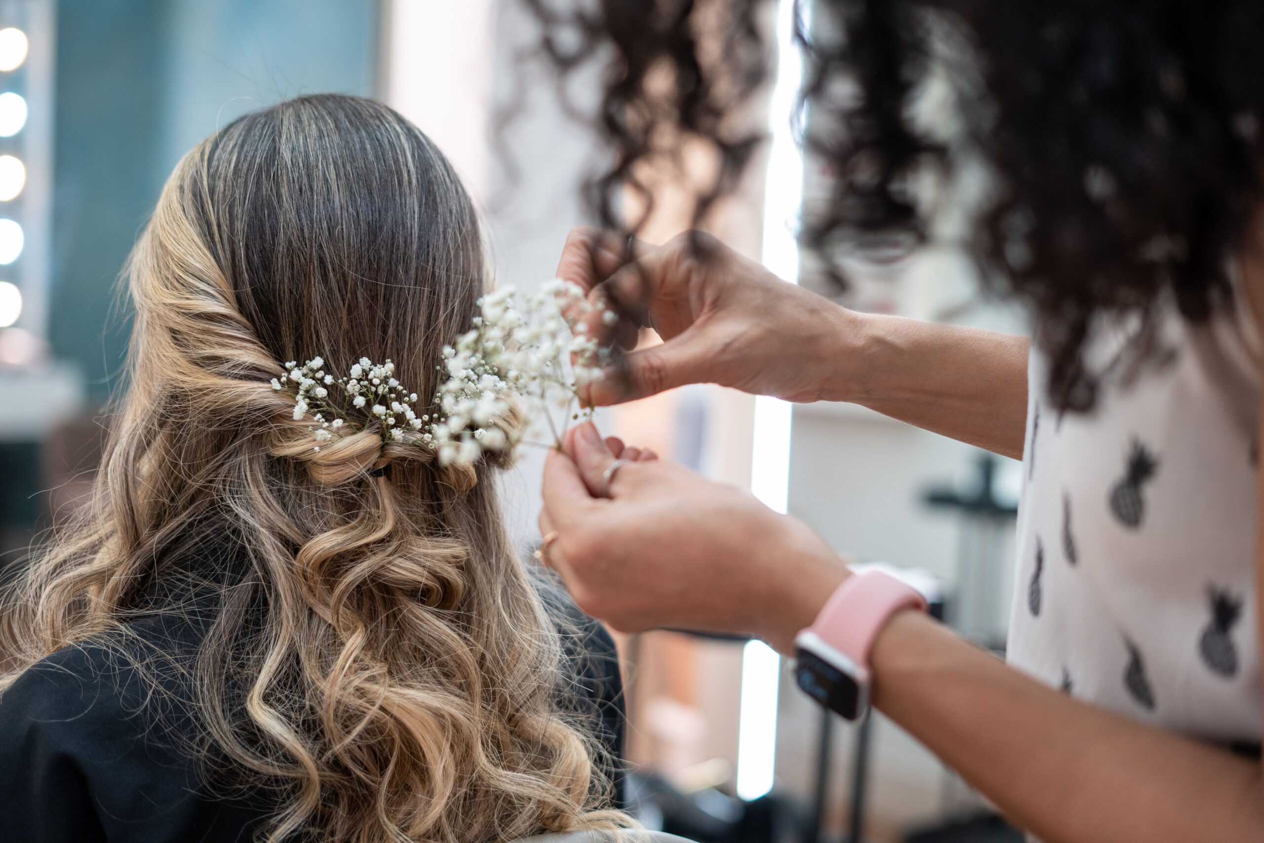 A hairstylist arranging delicate flowers in a woman's wavy hair, creating a bridal hairstyle in a salon setting.